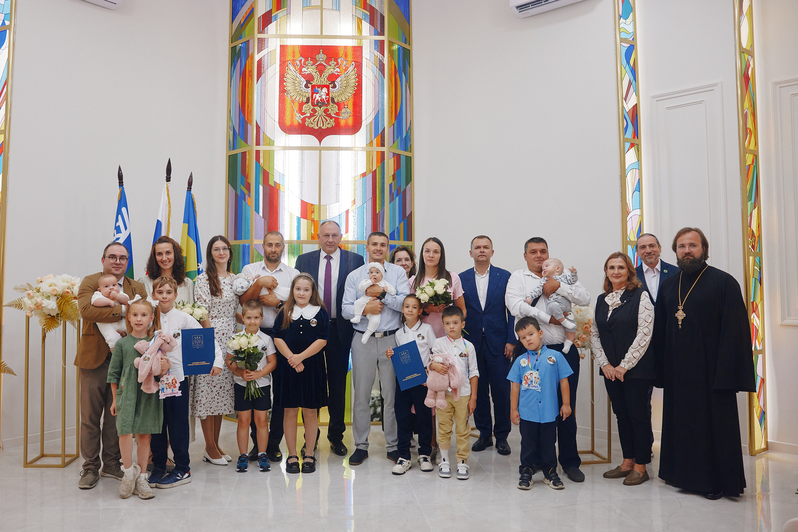 Фотография с поздравления многодетных семей Ханты-Мансийска. Все награжденные и приглашенные гости на одном фото. На заднем фоне влаги Российской Федерации, и ХМАО-Югры. большое витражное окно, букет из цветов.