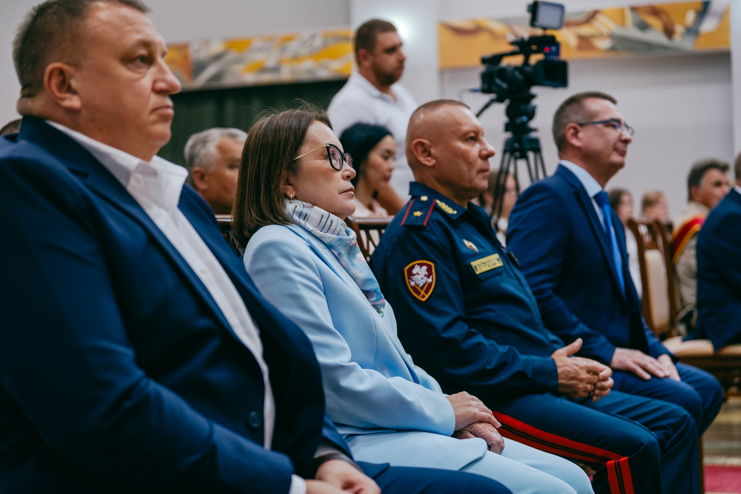В зале — зрители в первом ряду. Слева мужчина в синем костюме, рядом женщина в светло-голубом с очками, затем мужчина в военной форме с погонами и наградами, справа ещё один мужчина в костюме. Все смотрят в одну сторону — на сцену. На заднем плане — опера