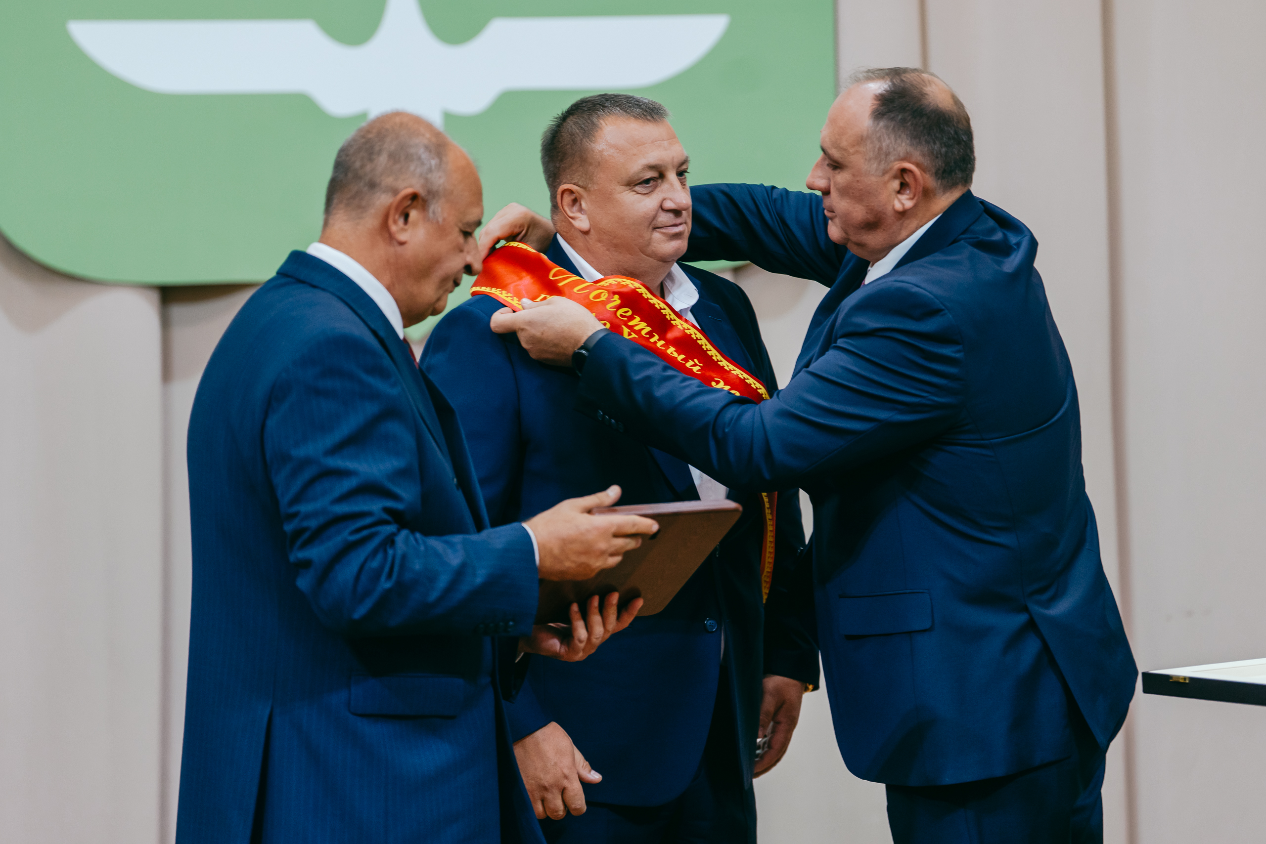 Трое мужчин в синих костюмах на сцене. Двое — слева и справа — надевают красную ленту через плечо мужчине в центре. В руках у одного из них — папка с наградой. За ними — зелёный баннер с белой птицей. Момент торжественный и тёплый.
