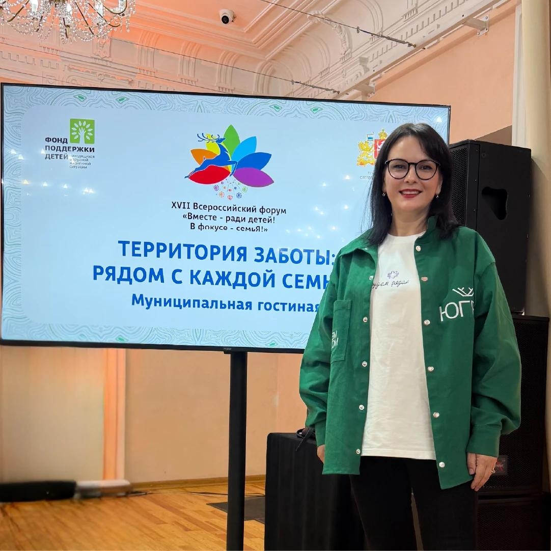 Женщина в очках и зелёной куртке с логотипом «Югра» стоит на фоне экрана. На экране — эмблема форума, надписи «Территория заботы: рядом с каждой семьёй» и «Муниципальная гостиная». За ней — колонна и зал с лепниной. Атмосфера деловая, официальная.