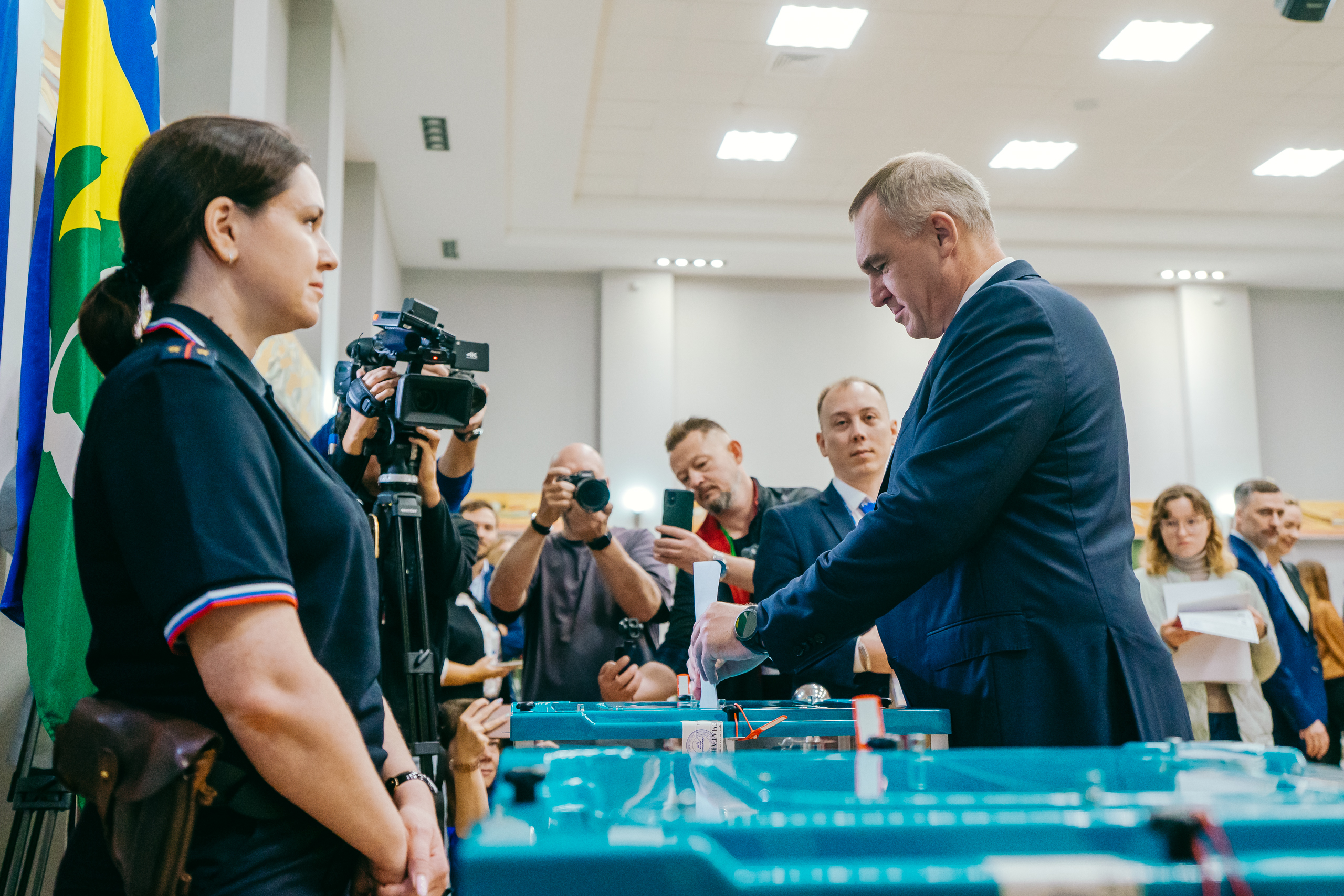 Мужчина в тёмно-синем костюме передаёт что-то женщине в форме с шевронами на рукаве. Они стоят у синего стола с оборудованием. Вокруг — операторы с камерами, фотографы и зрители. Слева виден флаг Югры. Атмосфера официальная, событийная.