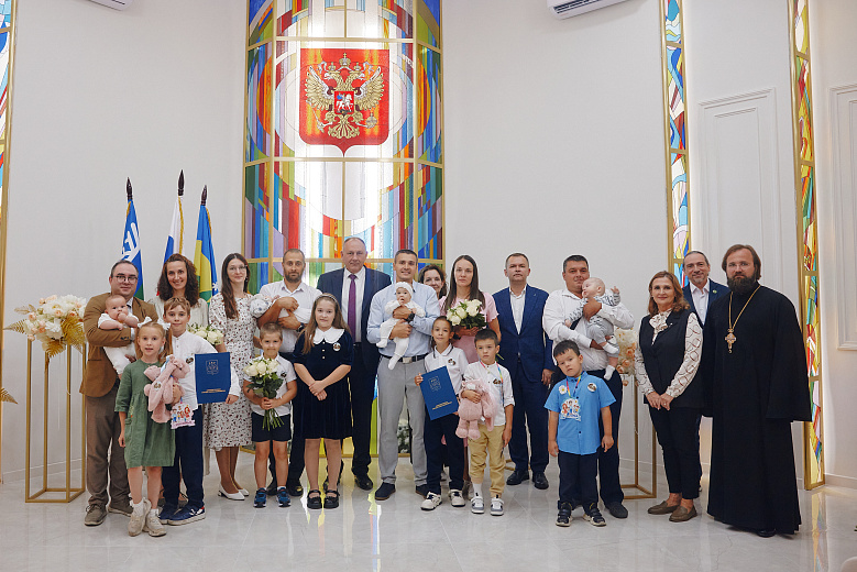 Фотография с поздравления многодетных семей Ханты-Мансийска. Все награжденные и приглашенные гости на одном фото. На заднем фоне влаги Российской Федерации, и ХМАО-Югры. большое витражное окно, букет из цветов.