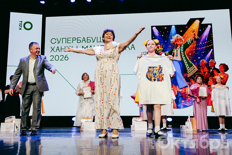 На сцене — женщина в светлом платье с цветочным узором, раскинув руки, улыбается. Рядом девушка держит белую футболку с принтом. Позади — ещё одна участница, а также декорация с изображением бабушки на фоне сцены.